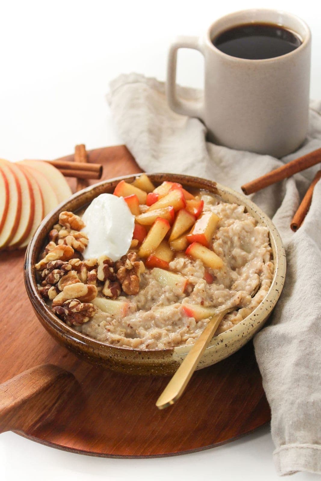A speckled ceramic bowl of apple cinnamon oatmeal topped with walnuts, a spoon of Greek yogurt, and diced apple, beside a mug of coffee.