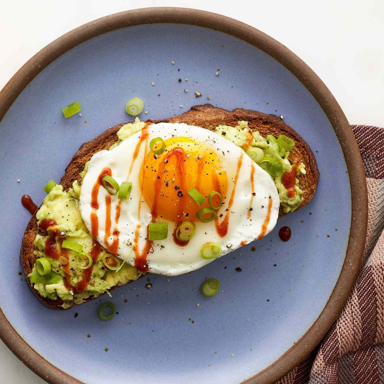 A fried egg with a bright yolk on smashed avocado toast, drizzled with harissa oil and scattered with sliced scallions.