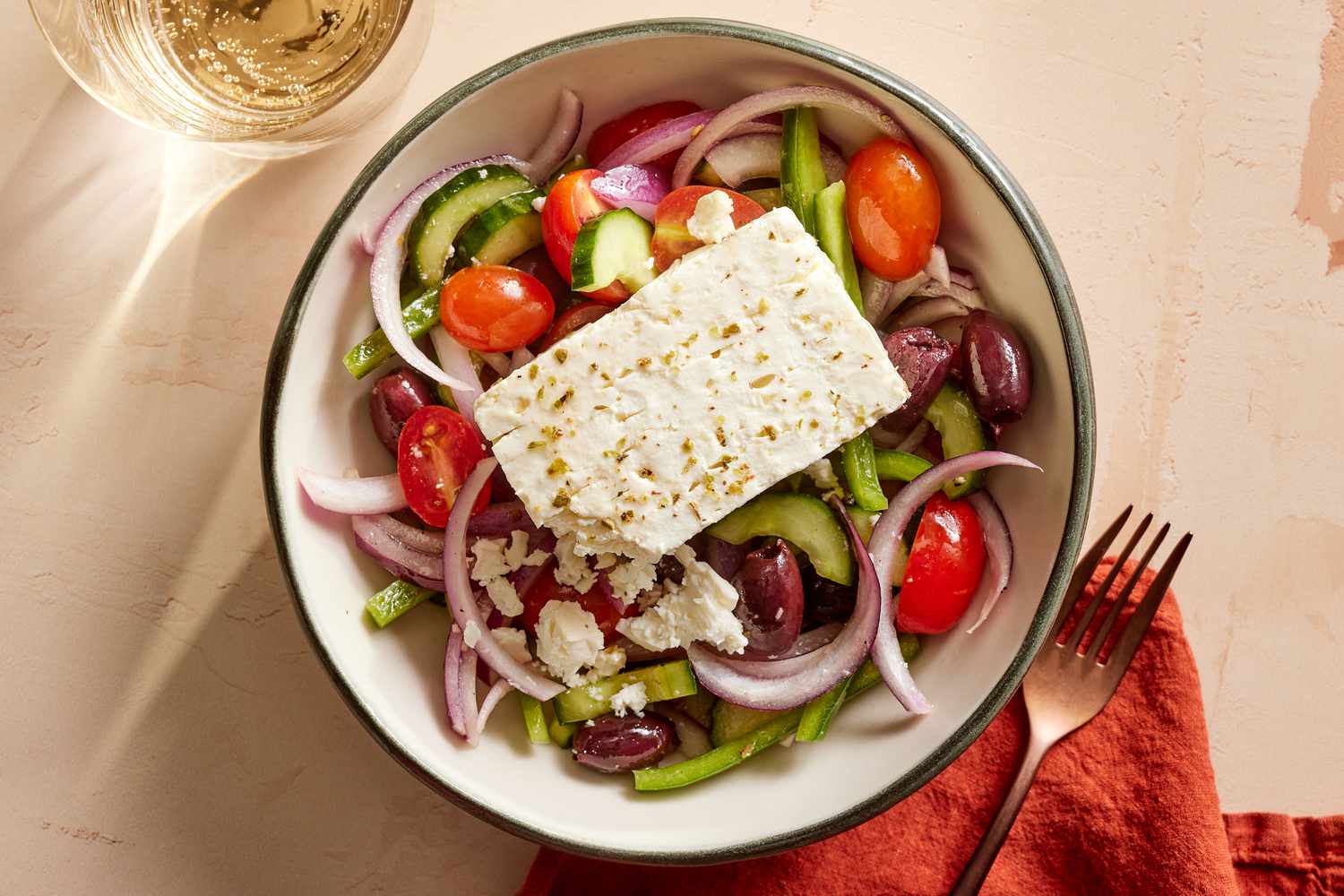 A bowl of Greek salad: tomatoes, cucumber, red onion, green pepper, and Kalamata olives under a slab of feta dusted with oregano.