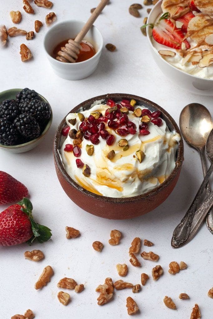 A small clay bowl of Greek yogurt topped with pomegranate seeds, chopped pistachios, and a swirl of honey, with berries and walnuts scattered nearby.
