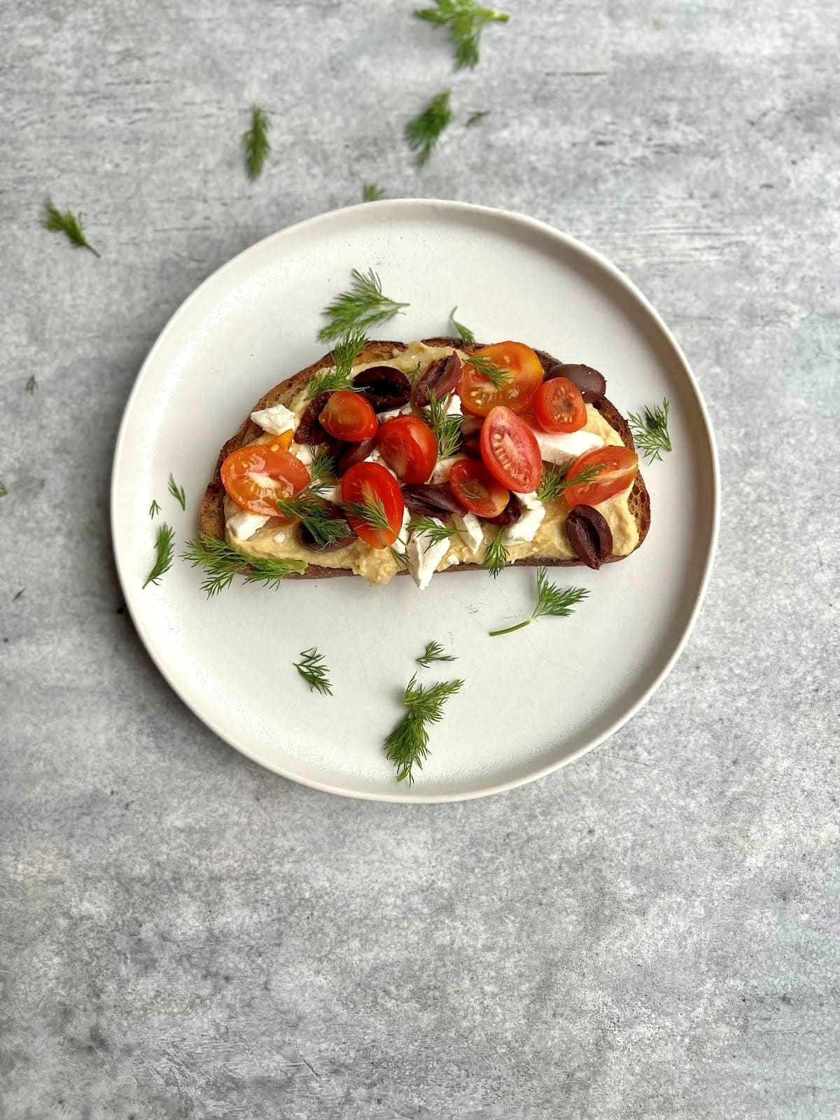 A slice of whole-grain toast spread with hummus and topped with halved cherry tomatoes, Kalamata olives, crumbled feta, and fresh dill.