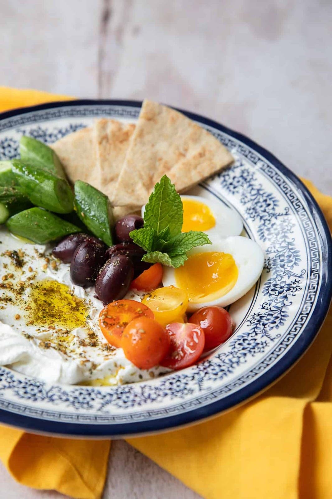 A blue-rimmed plate of labneh drizzled with olive oil and za'atar, with soft-boiled eggs, Kalamata olives, cucumber spears, cherry tomatoes, mint, and pita wedges.