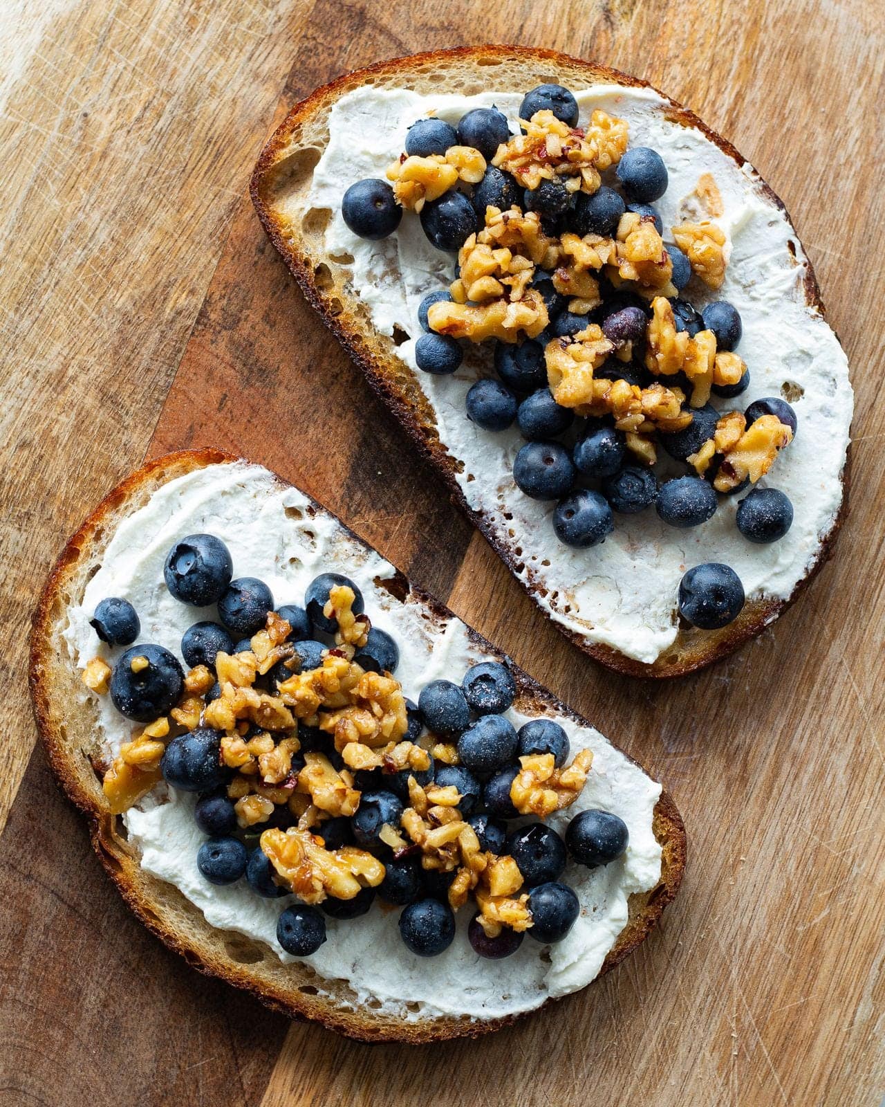 Two slices of sourdough spread with ricotta and piled with blueberries and honey-glazed walnuts on a wooden board.