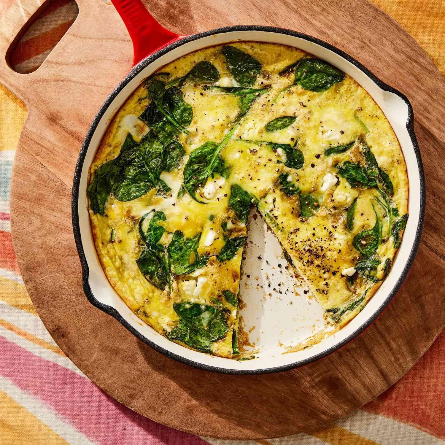 A spinach and feta frittata in a cast-iron skillet on a wooden board, with one wedge cut away.